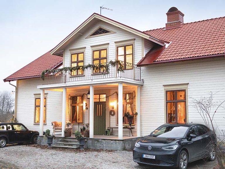 Beiget trähus med rött tegel, tända fasadlampor och två bilar parkerade framför hus